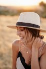 Mujer sonriente con cabello oscuro en sombrero y ropa elegante de pie con los ojos cerrados en el campo con hierba seca en el día soleado - foto de stock