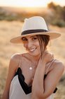 Mulher sorridente com cabelos escuros em chapéu e roupas elegantes em pé no campo com grama seca em dia ensolarado e olhando para a câmera — Fotografia de Stock