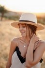 Mulher sorridente com cabelos escuros em chapéu e roupas elegantes em pé com olhos fechados no campo com grama seca no dia ensolarado — Fotografia de Stock