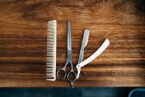 Top view of scissor and comb near straight razor with sharp metal blades on wooden table in hairdressing salon - foto de stock