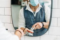 Crop anonymous male beauty master in sterile mask preparing shave brush with soap in bowl against mirror in bathroom at work - foto de stock