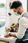 Side view of anonymous male beauty master in sterile mask preparing shave brush with soap in bowl against mirror in bathroom at work — Foto stock