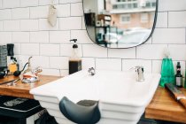 Table with assorted cosmetic products in bottles and dispensers between washstands under mirrors reflecting barbershop - foto de stock