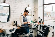 Вид сбоку мужской парикмахер просматривает интернет на нетбуке в современном салоне красоты напротив окна — стоковое фото
