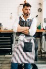 Self assured adult bearded male hairstylist in apron looking at camera with folded arms in barbershop — Stock Photo