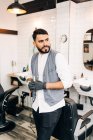 Самозабезпечений дорослий бородатий чоловічий перукар у жилеті, який дивиться, як стоїть в перукарні, тримаючи інструменти — стокове фото