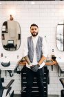 Self assured adult bearded male hairstylist in waistcoat looking at camera in barbershop - foto de stock