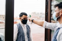 Crop anonymous male hairdresser in sterile mask measuring temperature of colleague with infrared thermometer at door of barbershop - foto de stock