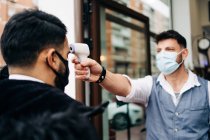 Male hairdresser in sterile mask measuring temperature of crop anonymous colleague with infrared thermometer at door of barbershop — Fotografia de Stock
