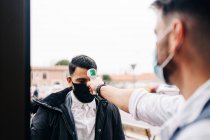 Crop anonymous male hairdresser in sterile mask measuring temperature of colleague with infrared thermometer at door of barbershop — Fotografia de Stock