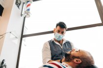 From below master barber on face medical mask trimming beard of masculine man with electric machine in hairdressing salon during covid pandemic — Fotografia de Stock