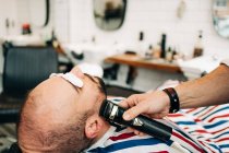 Crop anonymous master trimming beard of masculine man with electric machine in hairdressing salon — Fotografia de Stock