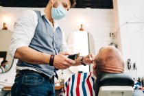 From below master barber on face medical mask trimming beard of masculine man with electric machine in hairdressing salon during covid pandemic — Stock Photo