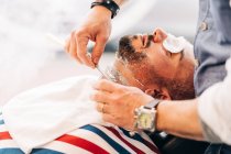 From above of crop anonymous beauty master in wristwatch shaving beard of client with straight razor during steam vapor treatment in hairdressing salon - foto de stock