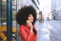 Vista lateral de la mujer afroamericana feliz con el pelo rizado sonriendo ampliamente mientras habla en el teléfono inteligente en la calle de la ciudad - foto de stock