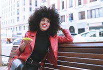 Elegante surpreendido afro-americano feminino lendo notícias no celular sentado no banco na cidade — Fotografia de Stock
