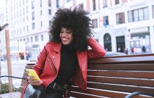 Стильный удивленный афроамериканка читает новости по мобильному телефону сидя на скамейке в городе — стоковое фото