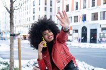 Side view of happy African American female with curly hair smiling widely while talking on smartphone on city street — Stock Photo