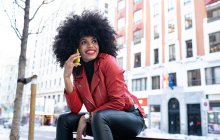 Side view of happy African American female with curly hair smiling widely while talking on smartphone on city street — Stock Photo