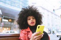 Elegante mujer afroamericana asombrada leyendo noticias en el teléfono celular sentado en el banco de la ciudad - foto de stock
