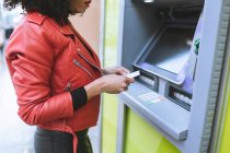 Donna africana americana anonima in piedi vicino al bancomat e che inserisce la carta di credito mentre sorride ampiamente e distoglie lo sguardo — Foto stock