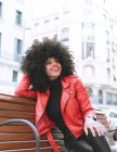 Low angle of cheerful African American female with Afro hairdo leaning on hand sitting with crossed legs on bench and looking away — Stock Photo