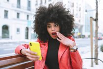 Elegante mujer afroamericana asombrada leyendo noticias en el teléfono celular con la boca abierta sentado en el banco de la ciudad - foto de stock