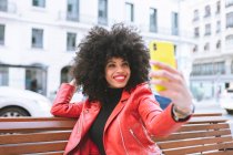 Вибіркове фокусування веселої афроамериканської жінки з африканською зачіскою сидячи на лавці і роблячи автопортрет на мобільному телефоні. — стокове фото