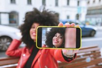 Enfoque selectivo de la mujer afroamericana alegre con peinado afro sentado en el banco y tomando autorretrato en el teléfono celular - foto de stock