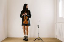 Cuerpo completo de violinista femenina de pie con instrumento musical y mirando cuidadosamente a la ventana - foto de stock