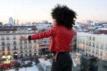Rückansicht einer Afroamerikanerin mit lockigem Haar im stylischen Outfit, die auf dem Balkon steht und auf städtische Gebäude blickt — Stockfoto