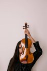 Künstlerin bedeckt Gesicht mit Geige, während sie vor weißem Hintergrund steht und in die Kamera schaut — Stockfoto