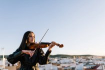 Молодая красивая женщина-музыкант в стильном мини-платье держит акустическую скрипку и стоит на крыше в жилом пригороде и смотрит в солнечный вечер — стоковое фото