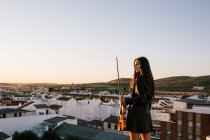 Voltar vista jovem bela músico feminino no elegante mini vestido segurando violino acústico e de pé no telhado em subúrbio residencial e olhando para longe na noite ensolarada — Fotografia de Stock