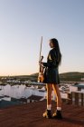 Back view young beautiful female musician in stylish mini dress holding acoustic violin and standing on rooftop in residential suburb and looking away on sunny evening — Stock Photo