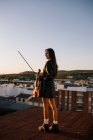 Смотреть назад молодая красивая женщина-музыкант в стильном мини-платье, держа акустическую скрипку и стоя на крыше в жилом пригороде и глядя в солнечный вечер — стоковое фото