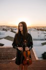 Junge schöne Musikerin in stylischem Minikleid mit akustischer Geige steht auf dem Dach eines Wohnviertels und schaut an einem sonnigen Abend weg — Stockfoto