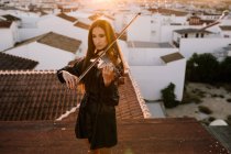 Jovem bela músico feminino em elegante mini vestido segurando violino acústico e de pé no telhado em subúrbio residencial e olhando para longe na noite ensolarada — Fotografia de Stock