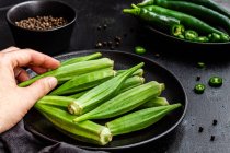Ernte unkenntliche Person hält reife Okra über Tisch mit frischem Gemüse auf Pfanne — Stockfoto