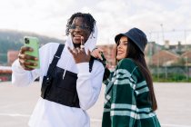Giovani partner contenuti multirazziali in abbigliamento elegante prendendo selfie sul marciapiede urbano alla luce del sole — Foto stock