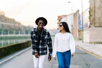 Jeunes partenaires multiraciaux de contenu en vêtements à la mode et lunettes de soleil parler tout en se regardant et se promener sur la chaussée de la ville — Photo de stock