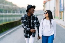 Young content multiracial partners in trendy clothes and sunglasses talking while looking at each other and strolling on city roadway — Stock Photo