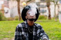 Serious African American male wearing stylish checkered shirt sunglasses and hat smoking cigarette — Fotografia de Stock