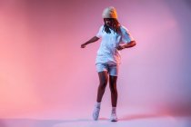 Corpo inteiro de adolescente concentrado afro-americano dançando com as mãos estendidas em estúdio com luz de néon brilhante — Fotografia de Stock