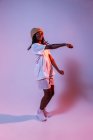 Corps complet de concentré afro-américain adolescent dansant avec les mains tendues en studio avec une lumière au néon brillante — Photo de stock