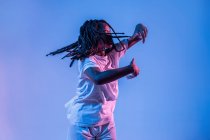 Dinâmica afro-americana adolescente fazendo movimento ao executar dança urbana em luz de néon contra fundo azul — Fotografia de Stock