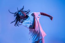 Dynamic African American teenage girl making movement while performing urban dance in neon light against blue background — Stock Photo
