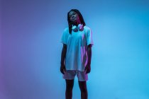 Chica adolescente afroamericana seria en auriculares en el cuello de pie en el estudio con luces de neón brillantes - foto de stock
