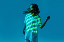 Dinámica afroamericana adolescente haciendo movimiento mientras realiza danza urbana en luz de neón sobre fondo azul - foto de stock