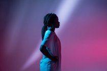 Side view of Serious African American teenage girl in headphones on neck standing in studio with bright glowing neon lights — Stock Photo